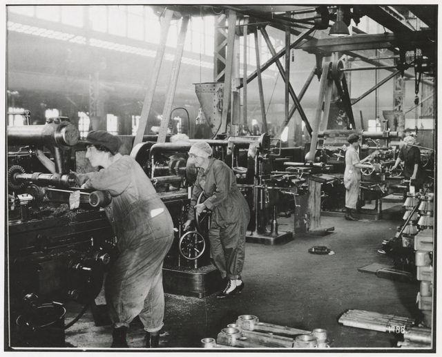 Two women in overalls stand in the left foreground, working with machines; two other women workers stand at right in the middle ground. Line shafts branch out above their heads.
