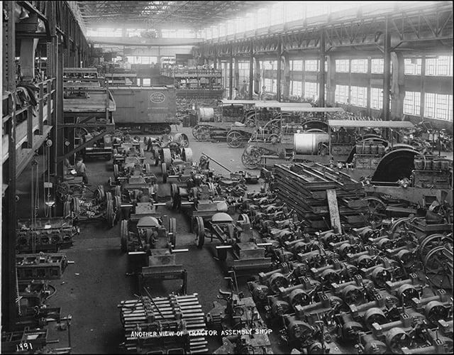 Interior view of a factory floor with dozens of tractors and machines stretching to a point in the background, with windows emitting light at the back and at the sides.