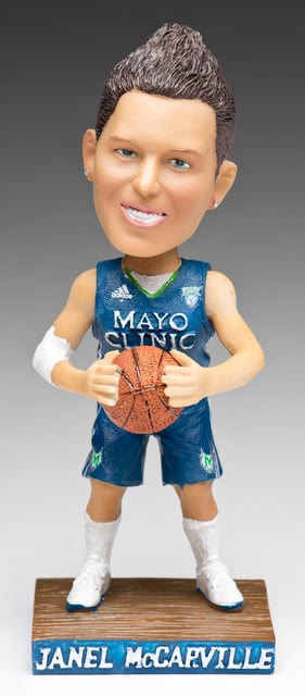 A bobblehead figure on a wood base against a background with a gradient of white to black. The figures wears a basketball jersey and holds a basketball.