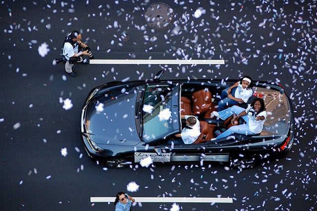 Two women, viewed from overhead, ride in a black convertible during a parade.