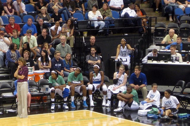 Corner of a basketball court mid-game, with fans seated in rows of tiered seats, and players sitting in chairs and on the floor below them. 