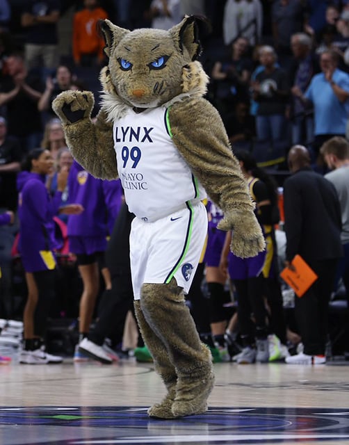 A full-body view of a basketball mascot in a lynx costume and white jersey, with players and fans in the background.