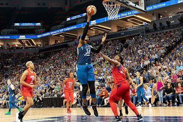 A basketball player in a blue uniform jumps toward a basket with a ball in hand. Three players wearing red uniforms surround her.