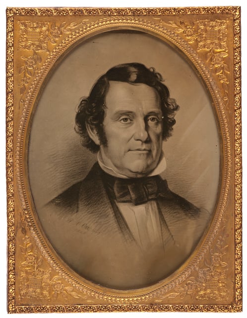 Gold frame with an oval black-and-white image of a man's head and shoulders. He wears a suit jacket with a bow tie and a high shirt collar.