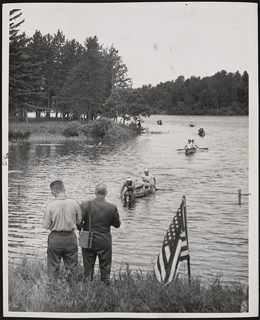 Two men stand on a river bank next to an American flag as canoists paddling in the water in front of them.