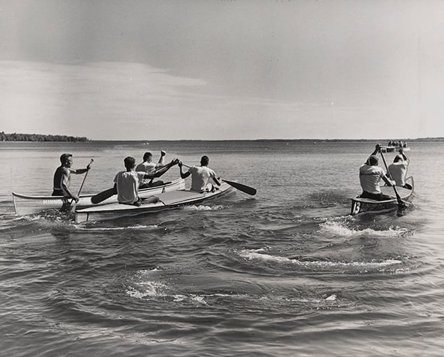 Rowers in three canoes paddle with oars in their hands across a lake in the middle ground, with the horizon and sky in the background.