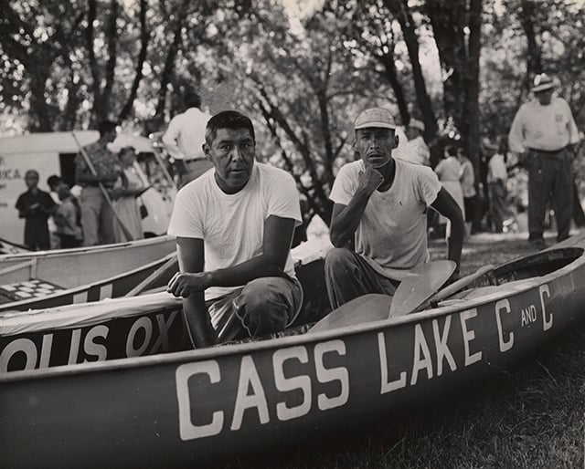 Two Ojibwe men crouch in a canoe with the words "Cass Lake" written on it in white capital letters.