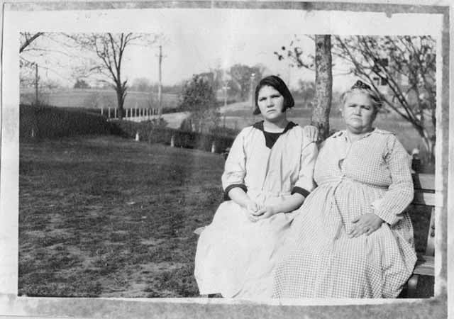 Two women sit on a bench in front of a field. The woman on the left wears a long white dress with a black collar and sleeve cuffs, and the woman on the right wears a long gingham dress