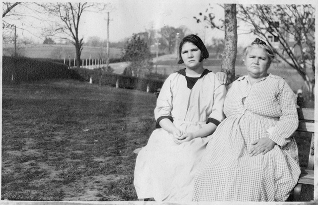 Two women sit on a bench in front of a field. The woman on the left wears a long white dress with a black collar and sleeve cuffs, and the woman on the right wears a long gingham dress.