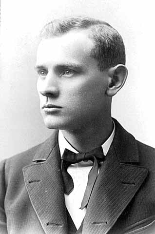 Photograph of 3/4 view of a man. He is wearing a blazer with a wide lapel, a ribbon bowtie, and a shirt with a white collar
