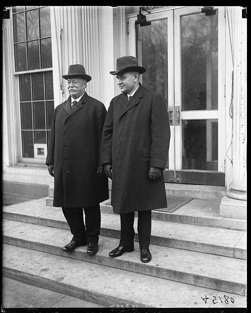 Two men in long dark coats stand on a set of stairs in front of a clear door.