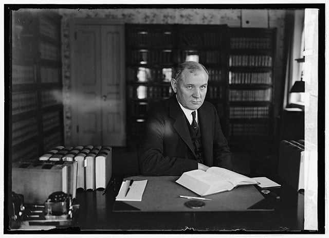 Three-quarter view of a man sitting at a desk. He wears a dark suit and tie. There are several closed books, a notepad, a pen, and an open book in front of him.