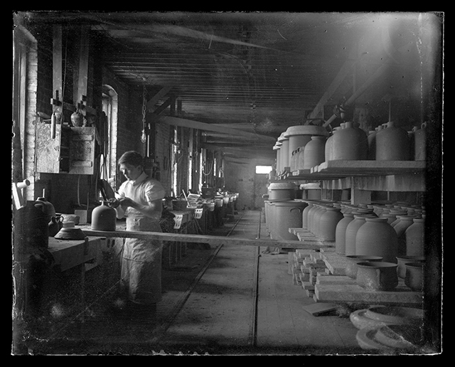 A worker stands inside a pottery storage room with windows on the left and ceramic vessels on the right, and a track leading through the center of the image into the backgroundd.