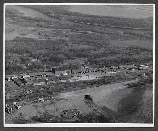 A bird's-eye view of a sprawling factory with buildings arranged into a narrow rectangle, with wooded land in the background.