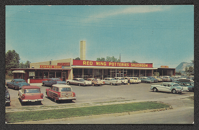 1950s-style cars sit in a parking lot in front of a single-story building with the words "Red Wing Potteries Salesroom" painted on the front facade.