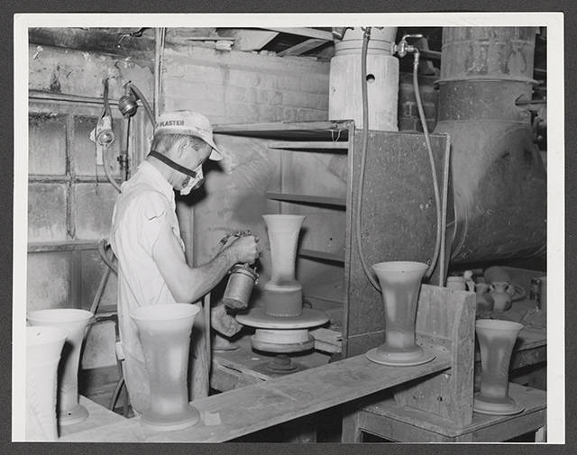 A worker wearing a brimmed cap and a respirator mask sprays glaze on a ceramic vase.