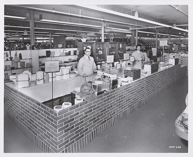 Two workers stand at sales counters behind a faux-brick wall.