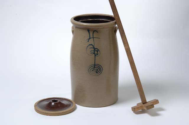 A cylindrical, narrow-lipped vessel stands against a gray background with a lid to the left and a wooden stick to the right.