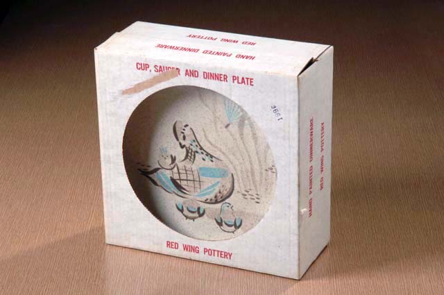 A ceramic plate is visible through an opening in a white box with red lettering against a coral background.