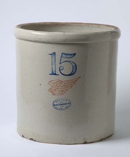 A dove-gray ceramic cylinder with the numeral 15 and a red wing image stamped on it sits in front of a lighter gray background.