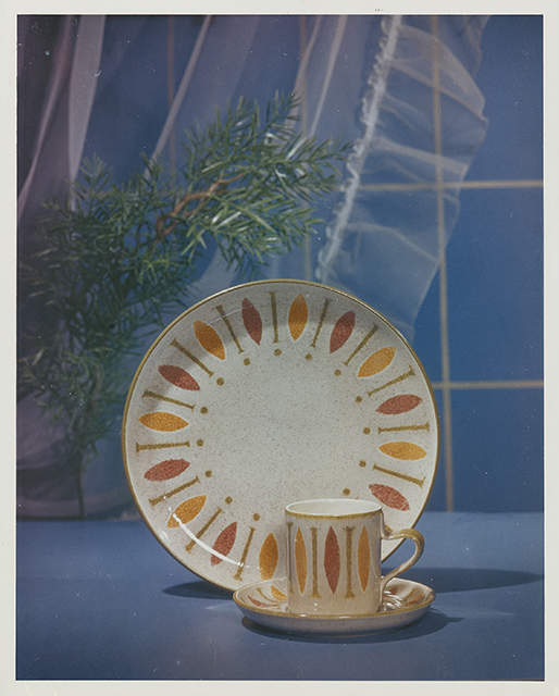 Ceramic plate and mug on a saucer with red and orange ovals on a blue surface with a white drape and green foliage in the background.
