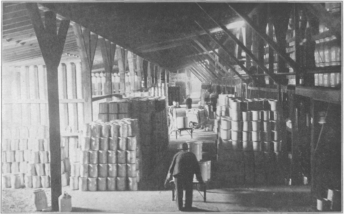 A worker in the foreground pushes a wheelbarrow down an aisle that leads between pyramidal stacks of ceramic containers.