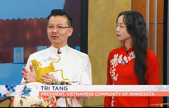 A man wearing Vietnamese traditional dress (áo dài) gestures with his hands and stands beside a woman also wearing áo dài. A screen a the wall of a TV studio are behind them.