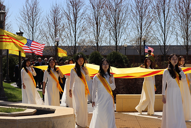 Nine women (visible) in long white dresses with yellow and red sashes carry a yellow and red flag.