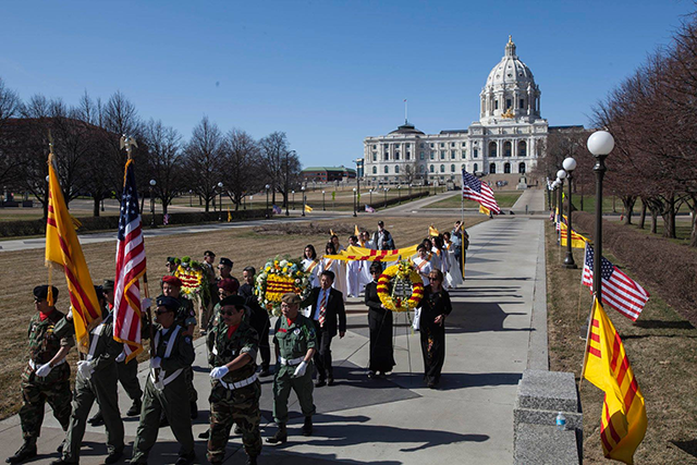Two rows and two clumns of people march on the Minnesota state capitol grounds carrying wreaths and flags. The capitol rises in the background against a blue sky.