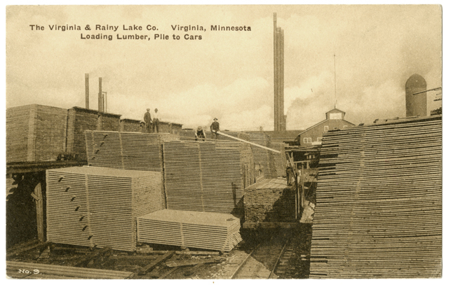 Stacks of wooden boards of varying heights sit together in a lumberyard, with an elevated track to the right.