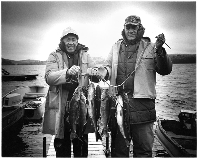 Two men stand side-by-side on a boat dock to hold up fish strung onto a line.
