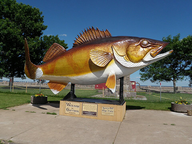 Yellow-and-brown sculpture of a fish with a white belly supported on a platform in front of trees and a blue sky.