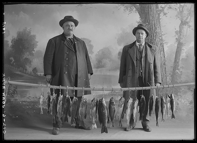 Two men in coats, hats, and suits stand in front of a line of walleye and perch in front of a photographer's scenic backdrop.