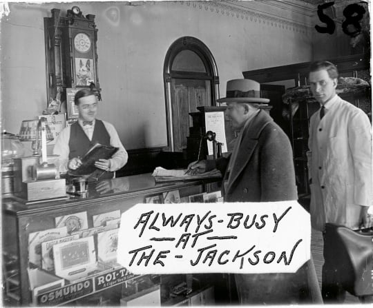 Image from a glass plate negative showing the front desk of the Jackson Hotel, ca. 1926. Cigars for sale are visible in the case, and a timetable for the Anoka Electric Line is fixed to the back of the cash register. Art Anderson is one of the men in the photograph. This image was used as an advertisement at Green’s Theater, located on Main Street in Anoka a few blocks from the hotel. Photographer unknown. Used with the permission of the Anoka County