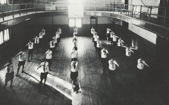 Black and white photograph of girls in uniform performing calisthenics during a physical education class on the second floor of Kiehle Hall, 1927.