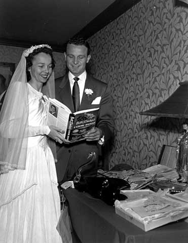 Black and white photograph of a wedding in the family of Mr. and Mrs. Frank Uram bride and groom at gift table looking at Betty Crocker cook book, 1950.