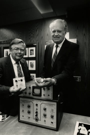 Black and white photograph of Medtronic Founder Earl Bakken and Medtronic CEO Winston Wallin with an early pacemaker model, 1986. Photographed by Jeffrey Grosscup.