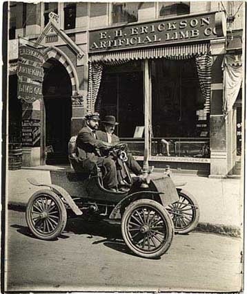 Black and white photograph of Michael Dowling and a passenger in a car in front of the E.H. Erickson Artificial Limb Company, c.1905.