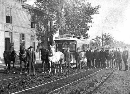 Black and white photograph of a horse-drawn streetcar in Minneapolis sign reads Sixth Street, Monroe Street and Eighth Avenue, c.1885. This photograph shows a second team of horses ready to relieve the team on the horsecar.