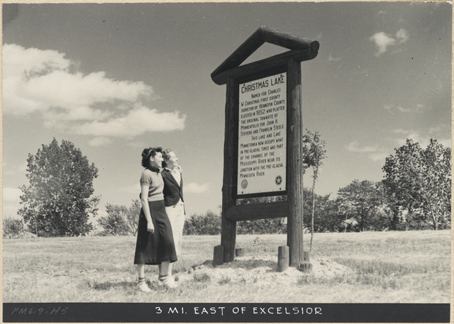 A pair of people stand before a white metal sign framed with a log mount. The marker is titled "Christmas Lake"