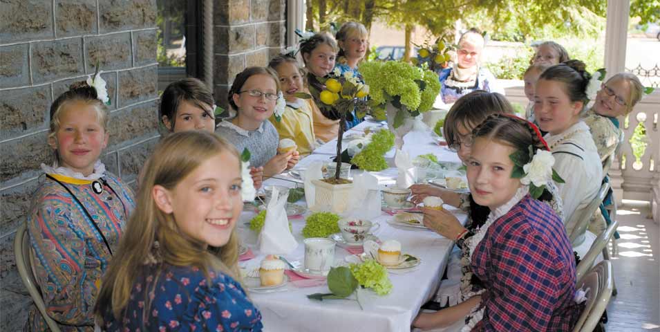 Girls in Victorian dresses sitting at a long table with tea and cupcakes
