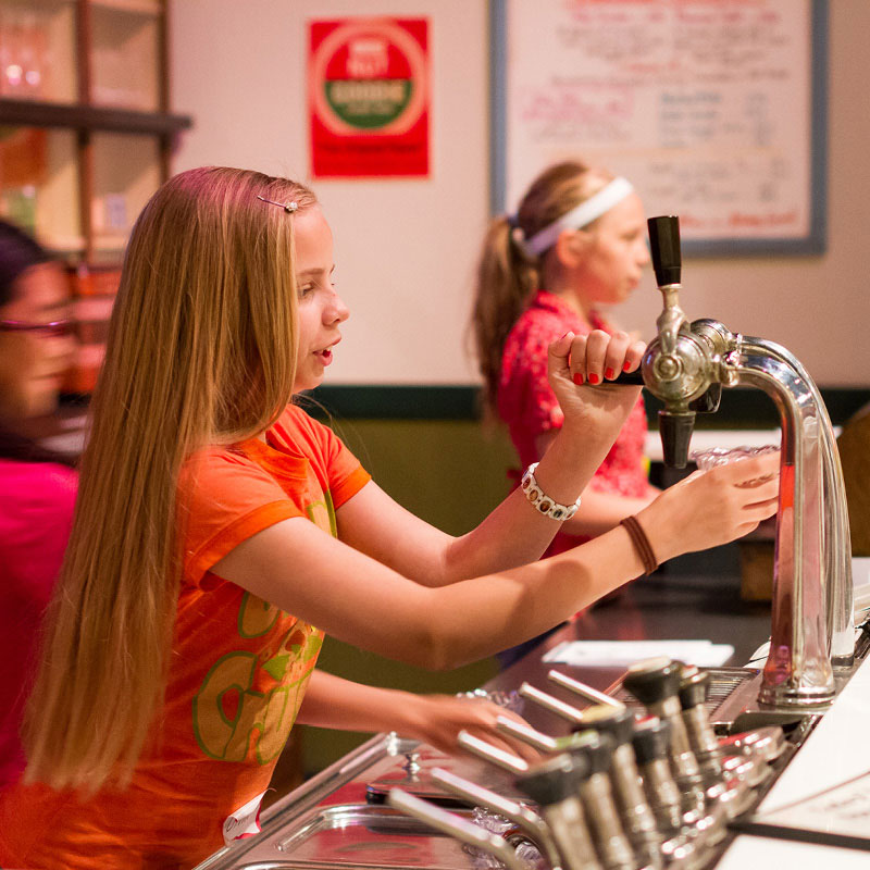 A girl pretending to pour soda from a 1930's era soda fountain.