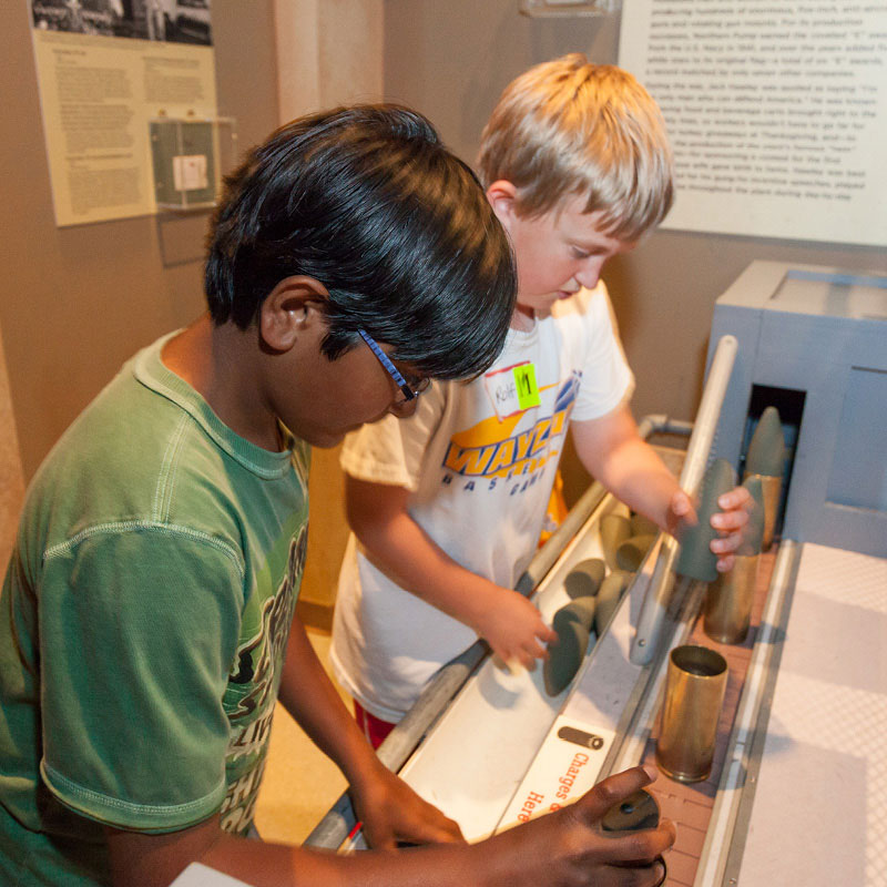 Two boys putting together shells in a bomb assembly line display.