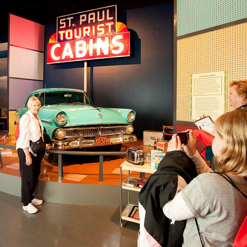 A woman standing in front of a 50's era car and a sign that says Saint Paul tourist cabins.