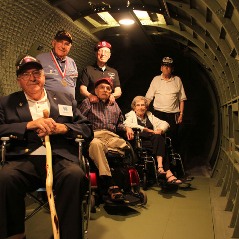 A group of military veterans standing and sitting inside the hull of an aircraft.