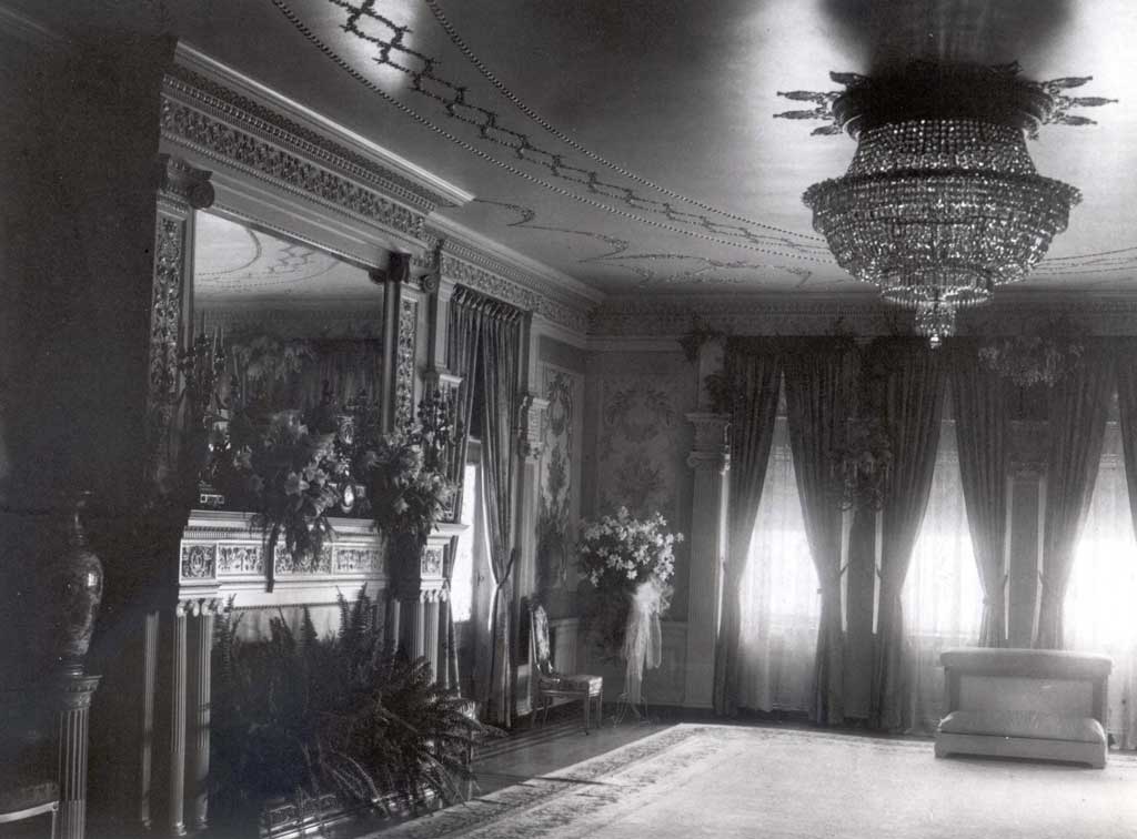 A room with sun coming through the windows, flower arrangements on the fireplace and in the corner