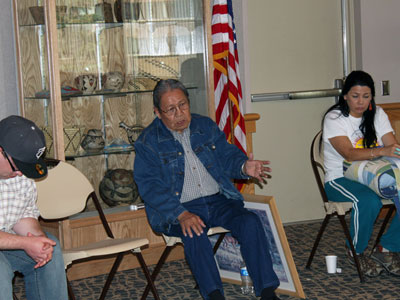 A seated Native American elder is talking with two other people seated nearby.