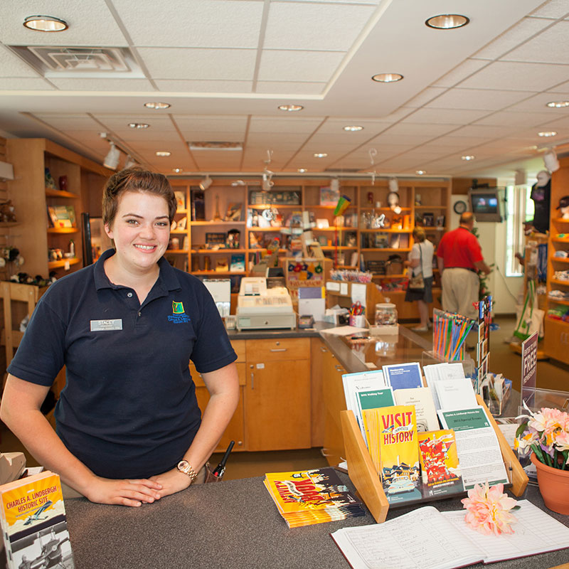 A store worker stands at the counter while several shoppers browse the store in the background.