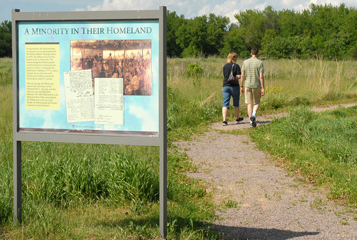 Two visitors walk outside on a dirt trail past an interpretive sign.