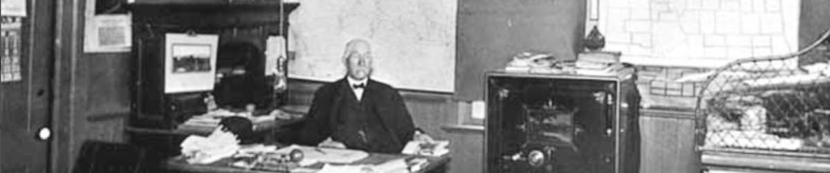An old man in suits sitting behind a desk, black and white photo.